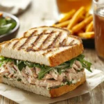 A grilled tuna salad sandwich with crispy bread, fresh greens, and side dishes on a rustic table.